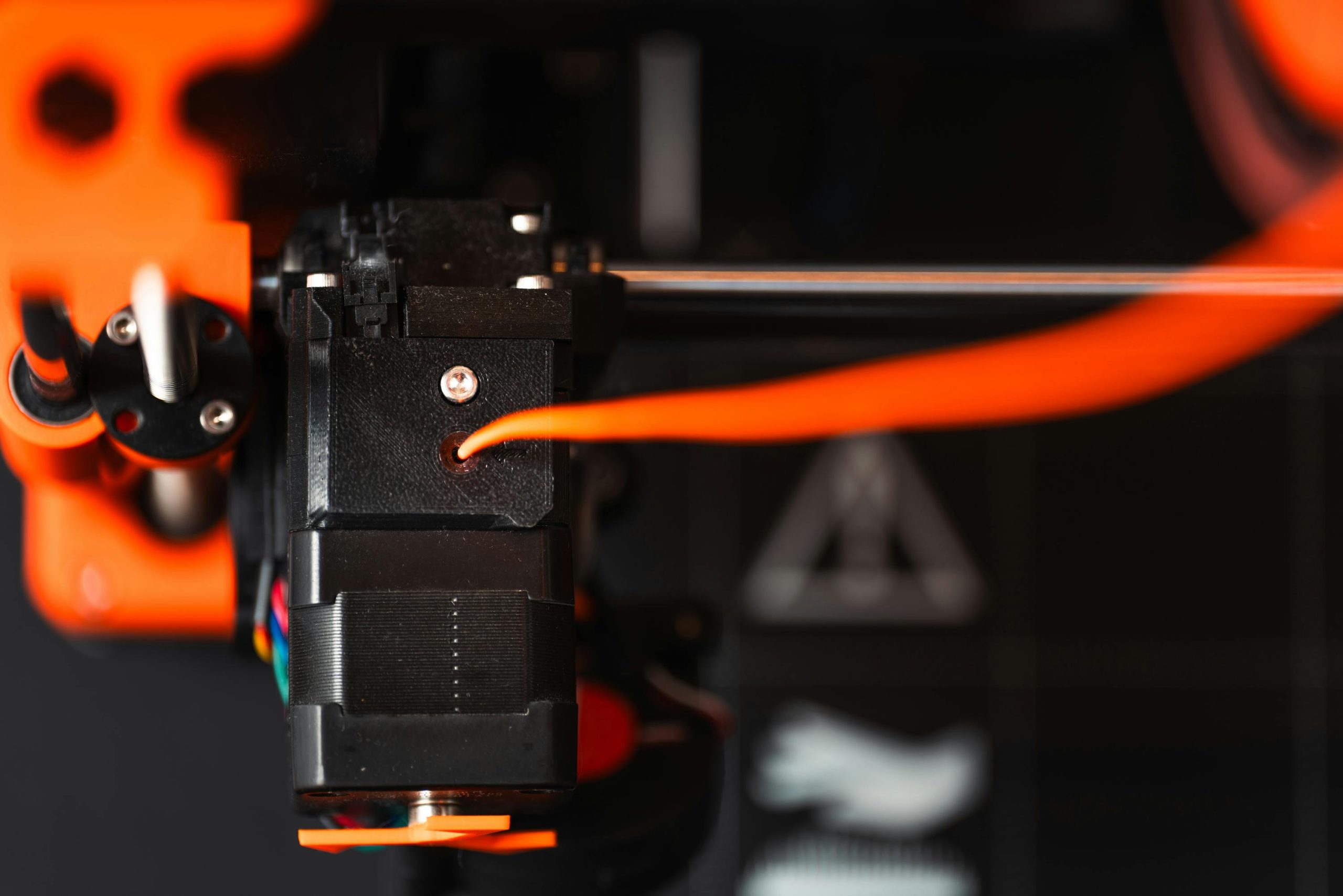Detailed view of a 3D printer extruder with orange filament in an industrial setting.